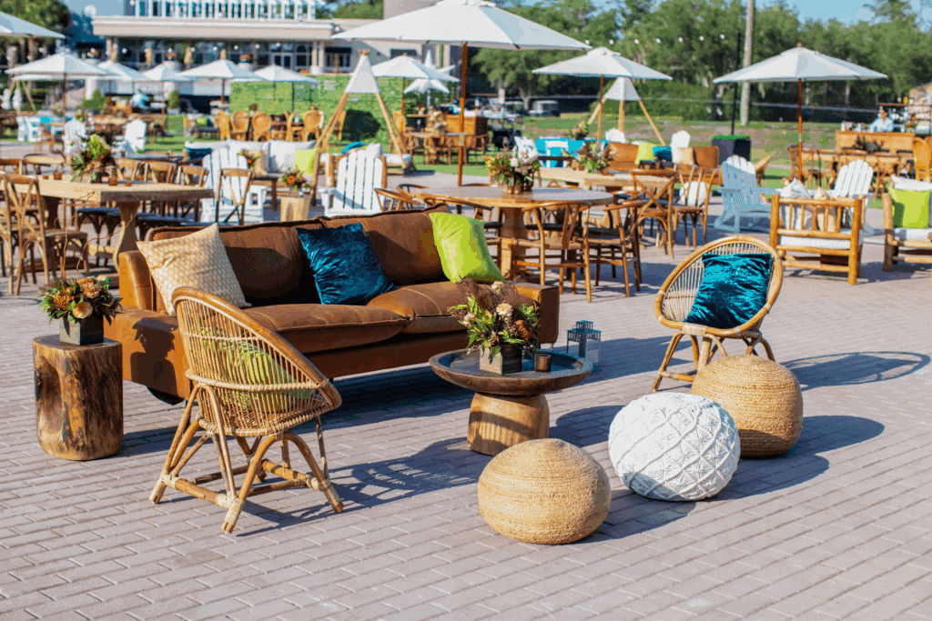 Outdoor corporate event lounge setup featuring mixed seating with modern and rustic furniture, vibrant accent pillows, florals, and umbrella-shaded dining areas — designed for guest comfort and elevated event aesthetics.